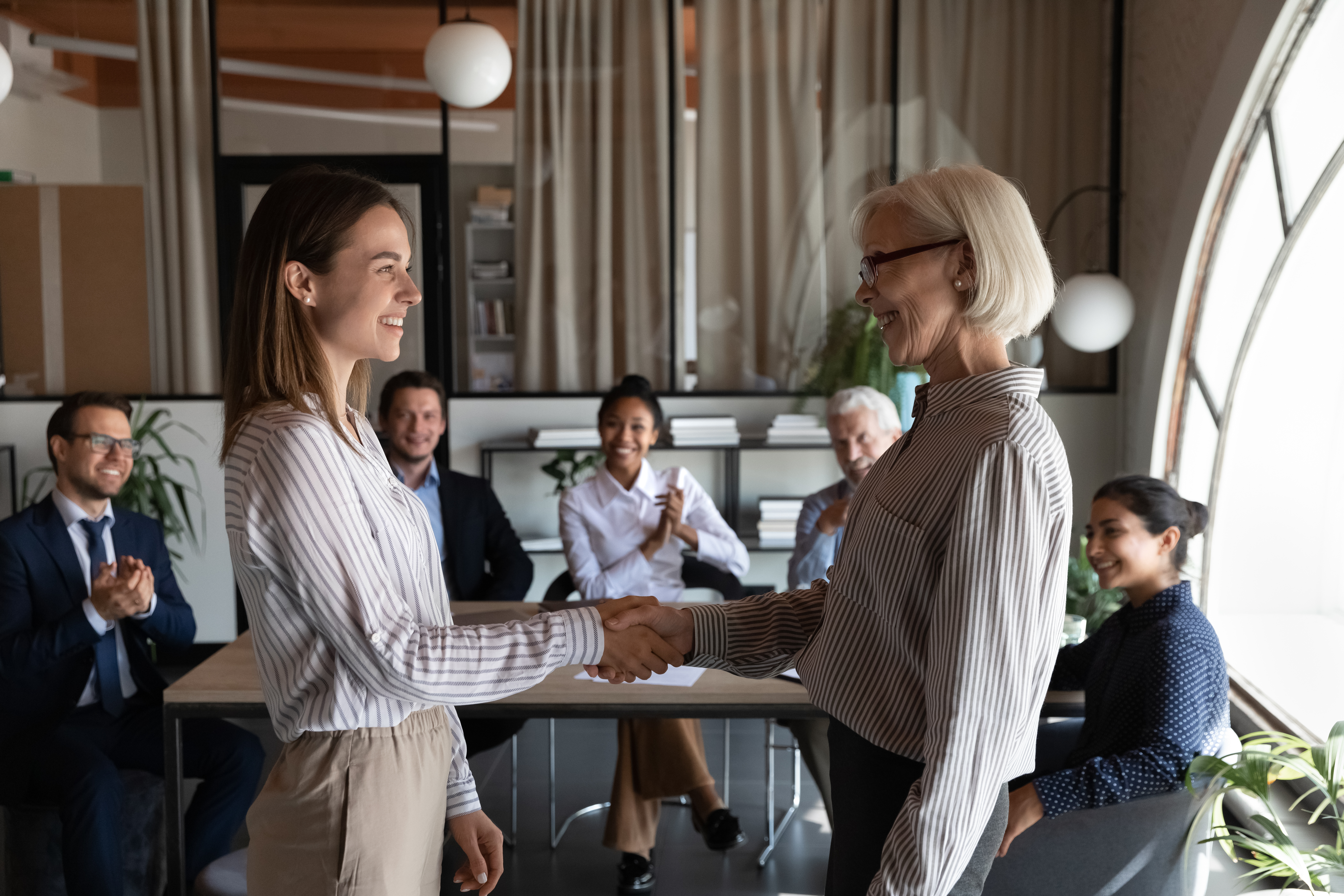 Photo of older-looking woman, white hair, lined face, shaking hands of younger-looking woman, both dressed similarly business casual, people at table clapping, one looking older, white hair, skin colors mixed -- white, brown, black.