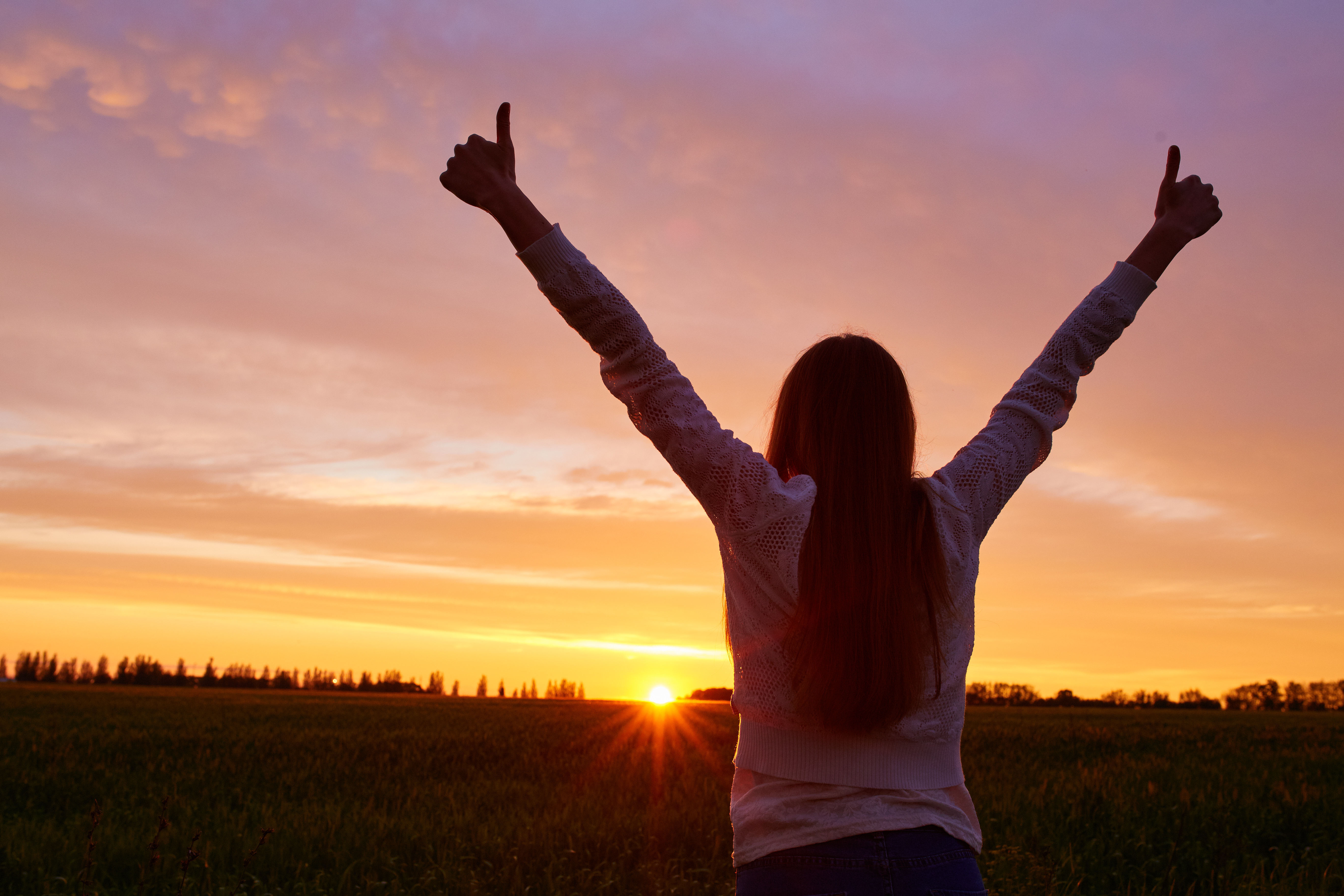 person with long hair (likely female), white skin, it appears, facing sun with thumbs up in the air