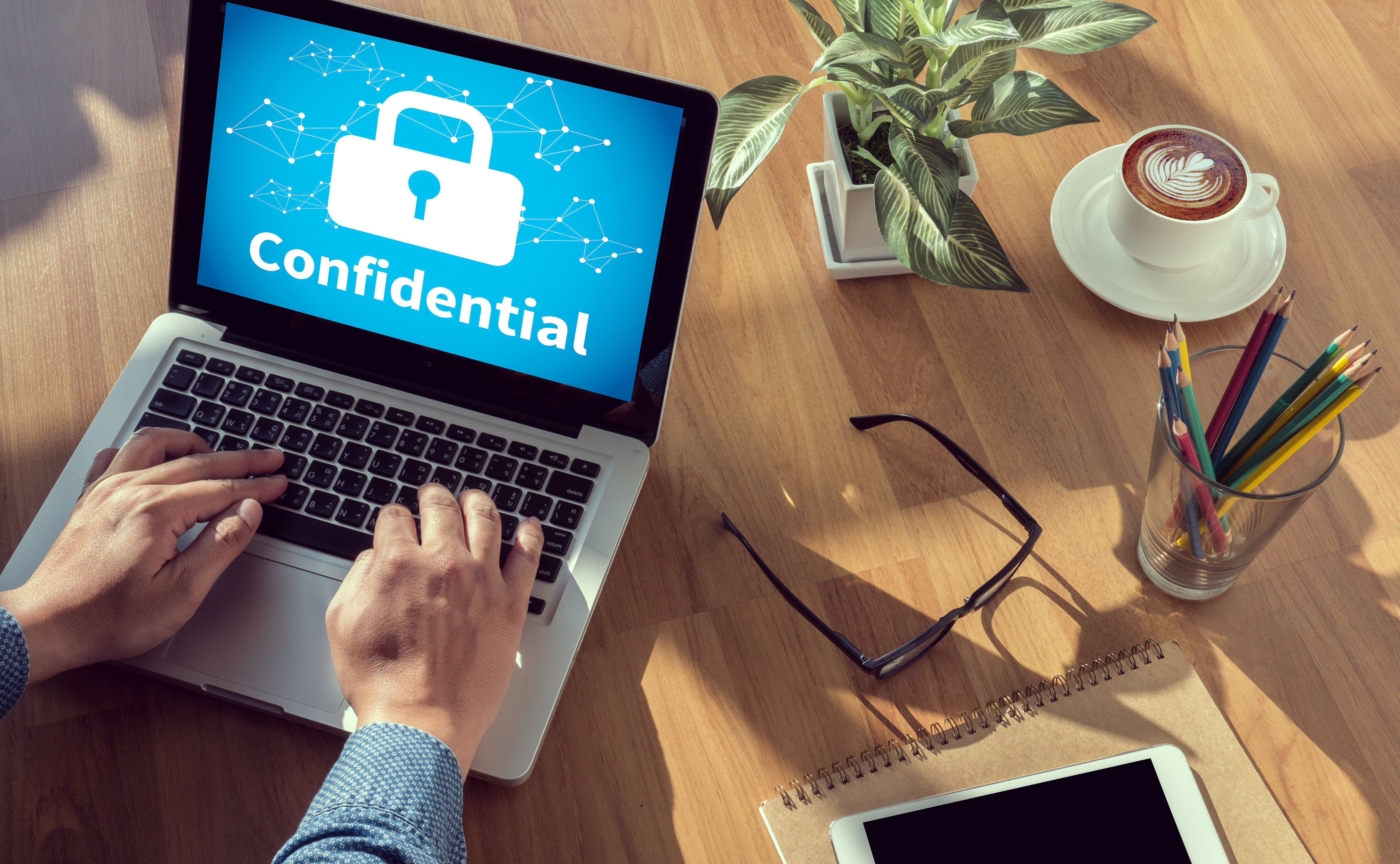 hands, appears white, working on laptop, screen says "confidential" and shows lock with key insert, other desk items shown -- coffee, glass with pencils, glasses, notebook, plant