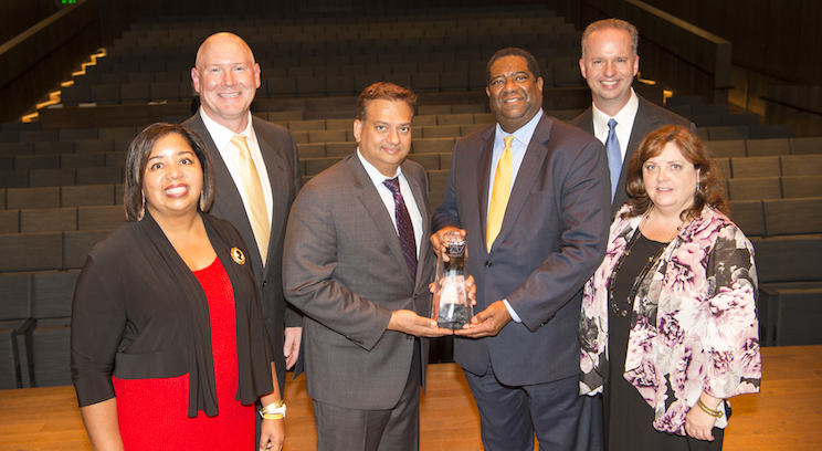a group of 6 people standing on stage. 2 people in the middle are holding an award