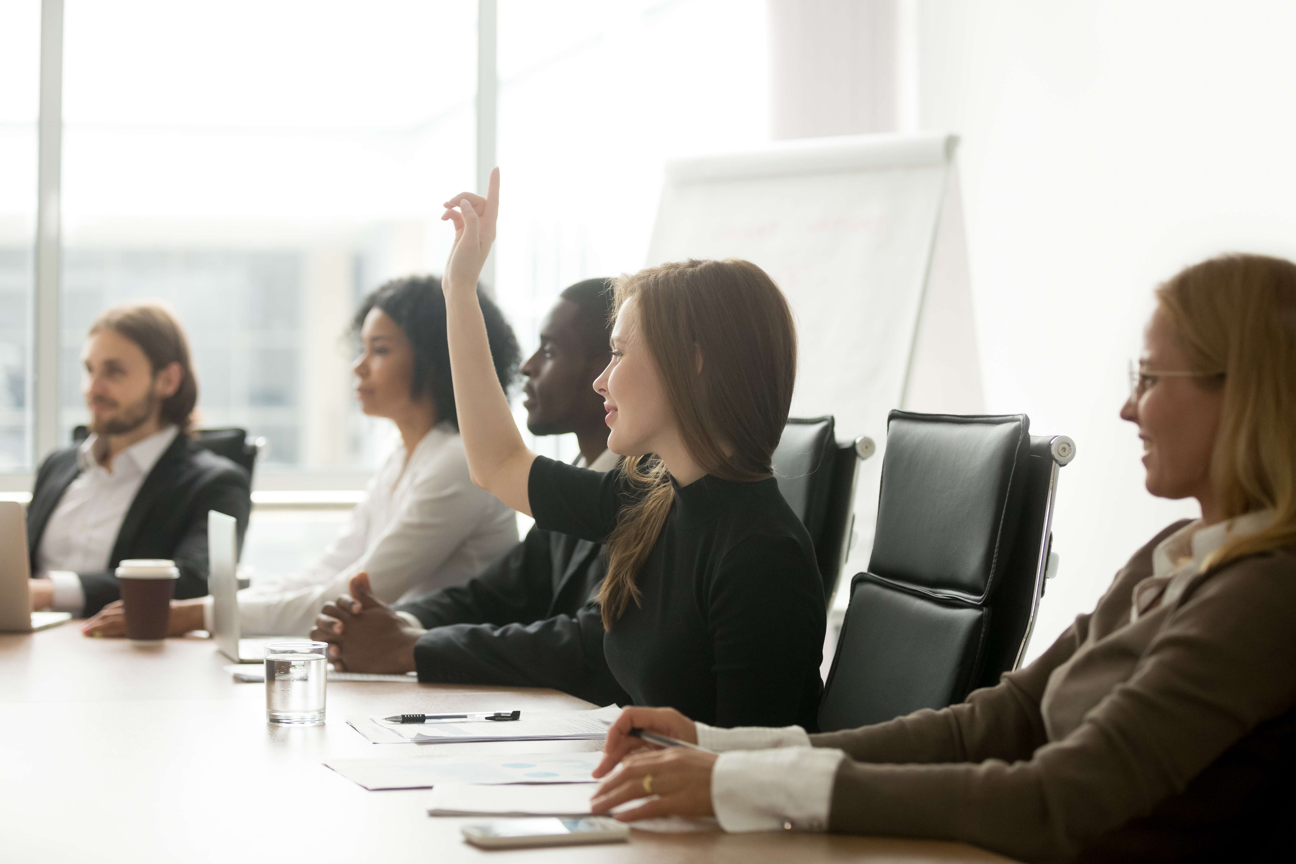 Smiling curious young businesswoman raising hand at group meeting.