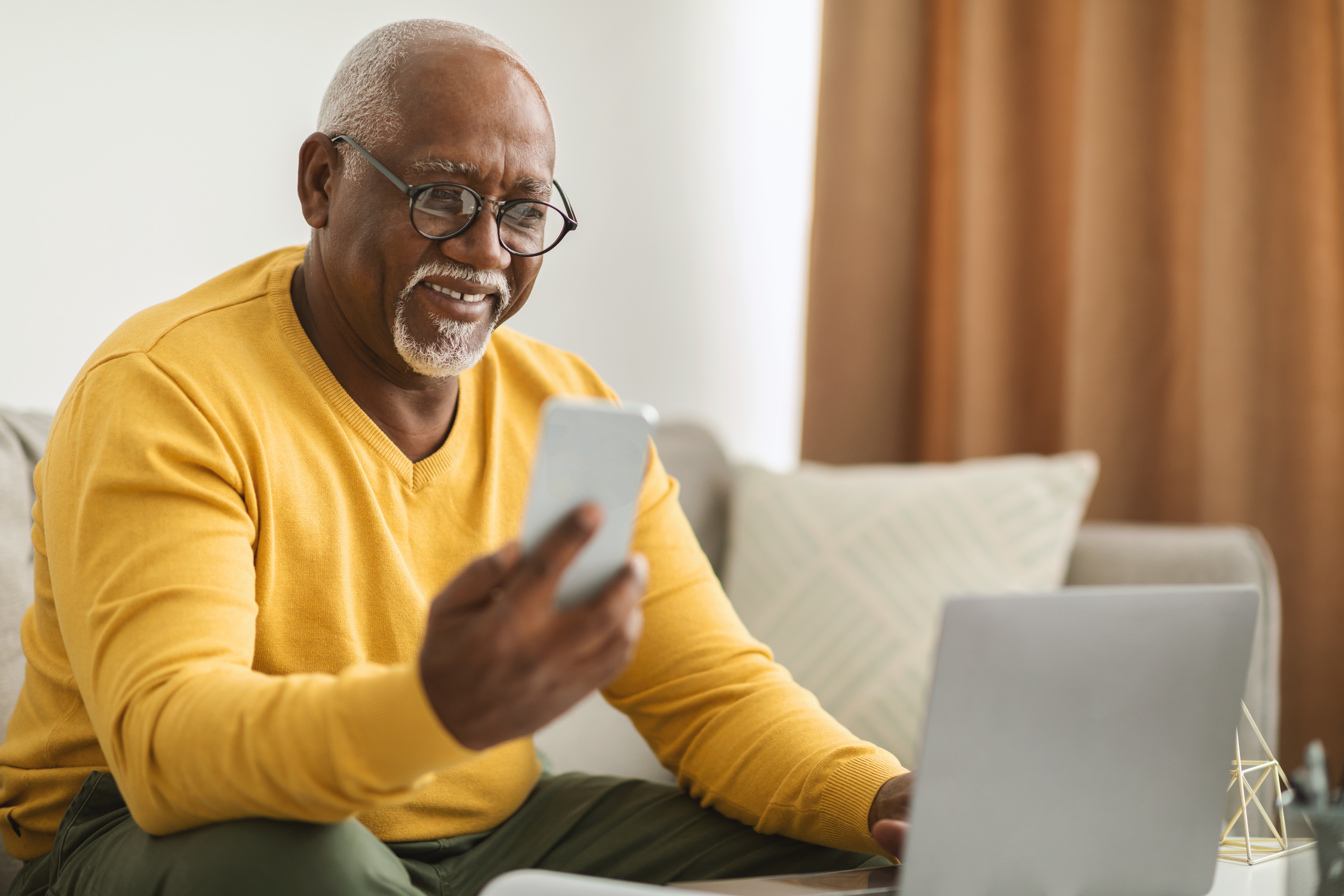 Photo of older-looking man, black-skinned, gray hair, multi-tasking with laptop and cell phone, business-casually dressed.