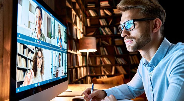 Man with glasses sitting in front of a teleconference in a dimly lit home library.