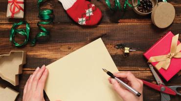 A hand poses with a pen to write on a blank piece of paper. On a table surrounded by Christmas wrapping materials.