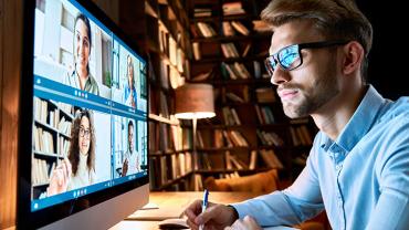 Man with glasses sitting in front of a teleconference in a dimly lit home library.