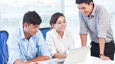 two professionals, a female and male employee sit at a desk looking at a computer while a third employee stands next to them