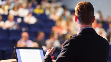 man at a podium, with back facing the viewer, speaks to an audience in the background
