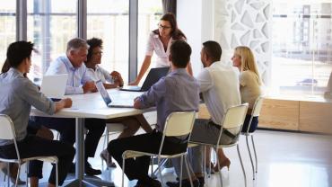 woman standing up in front of a table with her team seated around it, three on one side with three on the other side