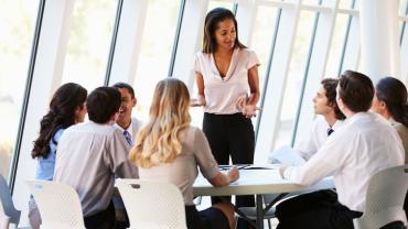 young professional woman standing up at a table with her team seated around her