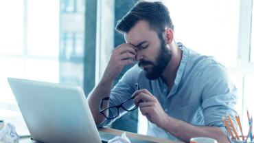 man seated at his computer with his glasses off holding the bridge of his nose