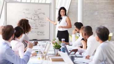 woman standing at a dry erase board giving a presentation to a group seated at the table