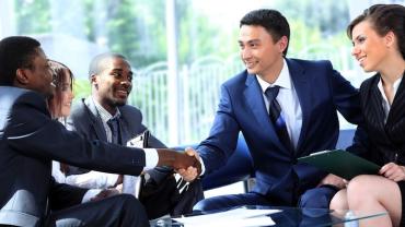 There are five people visible. a woman smiling on the far right. To her left is a man about to stand up shaking hands with another man seated across from him. In between these 2 men are a woman and another seated man all wearing business suits. 