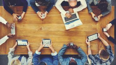 team of people sitting around a wooden table all using different technological devices