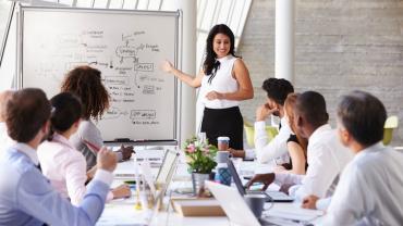 female leader standing next to dry erase board talking to her seven person team seated at a table