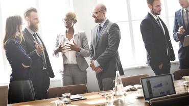 on the right a pair of businessmen talk and look at a tablet, while a group of four professionals stand next to them talking together during a meeting break