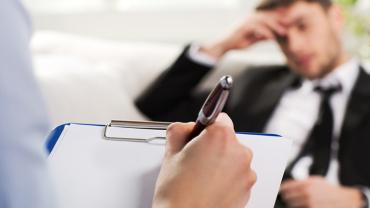 in the foreground is a hand holding a pen writing on a clipboard while in the background there is a male professional in a suit, tie is loose with his hand on his forehead