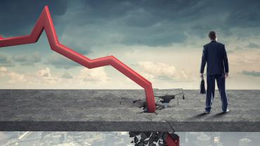 Red line chart plummeting into sidewalk next to a businessman in front of a cloudy gray sky.