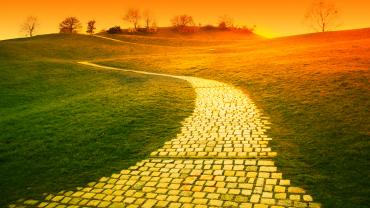Yellow brick road winding into the distance of a green field toward an orange sunset.