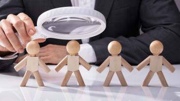 person in suit, white hand with magnifier examining wood figurines lined up like is staff needing investigation