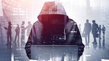 hooded figure, face not showing, hulking over laptop, figures and buildings in background, as though hooded one is hacking into their network