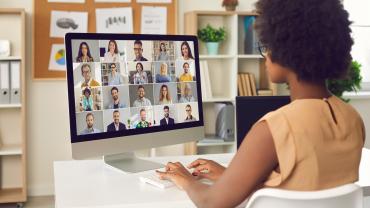 Black woman, appears to be, at desk with array of people on screen, far off but appears to be different skin colors