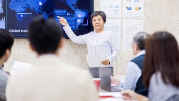 photo of, appears to be Asian woman, gray in hair, addressing group sitting, mixed ages, gender, culture, looking smartly business casually dressed, pointing at screen, smiling