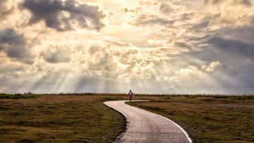 path curving with human figure in the distance, through grass toward sky with light beaming from clouds