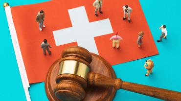 Large Swiss flag with gavel laying across it and small people standing around the gavel. 