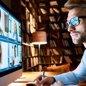Man with glasses sitting in front of a teleconference in a dimly lit home library.