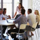 woman standing up in front of a table with her team seated around it, three on one side with three on the other side