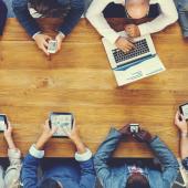 team of people sitting around a wooden table all using different technological devices