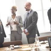 on the right a pair of businessmen talk and look at a tablet, while a group of four professionals stand next to them talking together during a meeting break