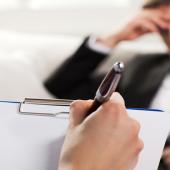 in the foreground is a hand holding a pen writing on a clipboard while in the background there is a male professional in a suit, tie is loose with his hand on his forehead