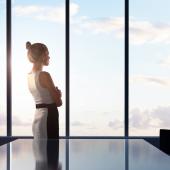 Businesswoman standing in front of a board room table looking out of a glass window in a modern office.