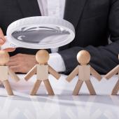 person in suit, white hand with magnifier examining wood figurines lined up like is staff needing investigation
