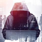 hooded figure, face not showing, hulking over laptop, figures and buildings in background, as though hooded one is hacking into their network