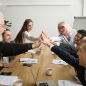 bunch of people gray-haired and not, seeming senior and young, at conference table, flip chart and plant in back, white, dark-photo
