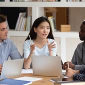 person talking around laptops with three people appearing to be listening intently, mixed genders and skin colors