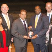 a group of 6 people standing on stage. 2 people in the middle are holding an award